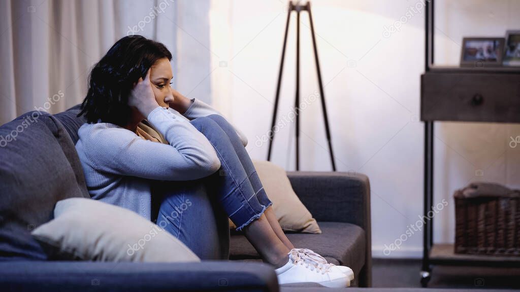 depressed woman in casual clothes sitting on sofa and holding hands near head at home