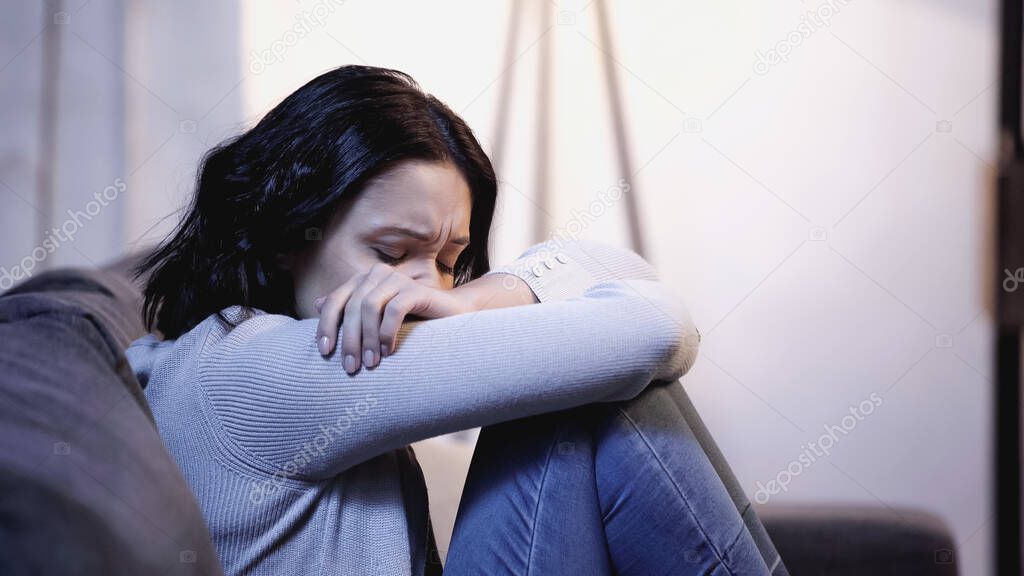 upset woman in casual clothes crying on sofa and hiding face in crossed arms at home