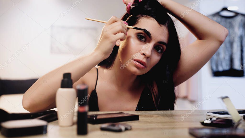 brunette young woman adjusting hair and styling eyebrow with brush near table with decorative cosmetics in bedroom