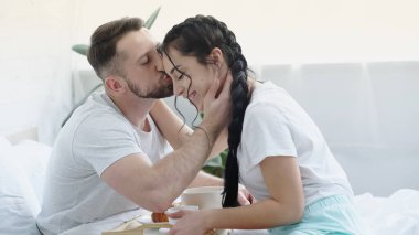 Sakallı adam, saçları örgülü mutlu esmer kadını öpüyor ve yatak odasına kahvaltı tepsisi getiriyor.