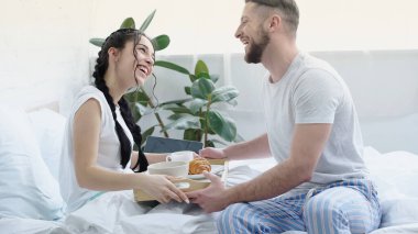 cheerful man bringing breakfast tray to smiling girlfriend with braids in bedroom  clipart
