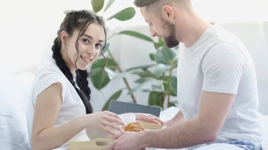 bearded man bringing breakfast to cheerful girlfriend with braids in bed clipart