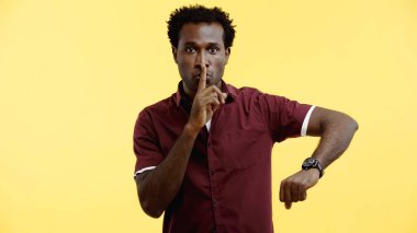 curly african american man in burgundy shirt and wristwatch showing hush sign isolated on yellow clipart