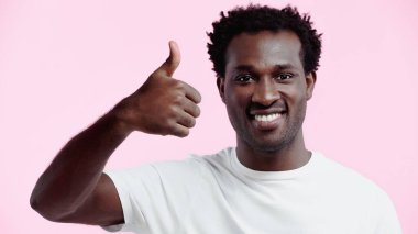 Beyaz tişörtlü, mutlu Afro-Amerikan adam pembe tişörtlü, parmaklarını gösterirken kameraya bakıyor.