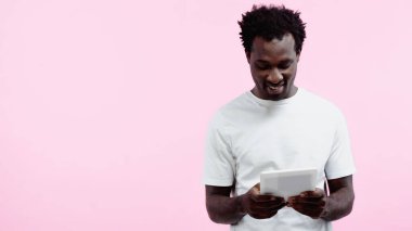 Beyaz tişörtlü neşeli Afro-Amerikan adam pembe tablet kullanıyor.
