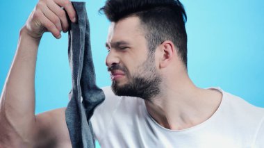 grimacing man smelling stinky sock isolated on blue clipart