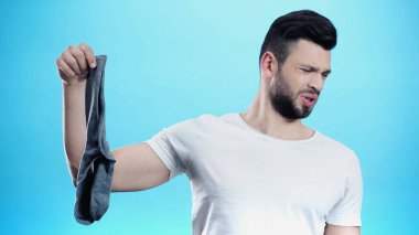 man feeling disgusted while holding smelly sock isolated on blue clipart