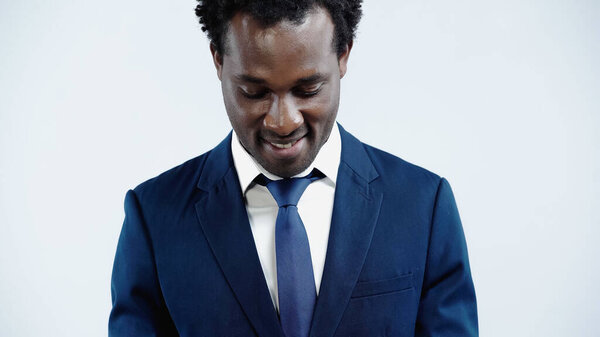 счастливый африканский американский бизнесмен в костюме с галстуком глядя вниз изолированы на синий