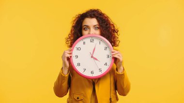 curly woman covering face while holding clock isolated on yellow clipart