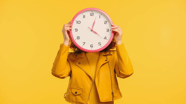 young woman covering face while holding clock isolated on yellow