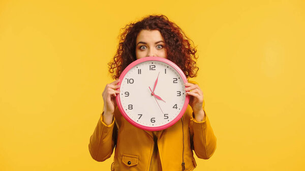 curly woman covering face while holding clock isolated on yellow