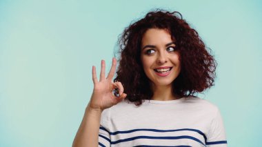 cheerful young woman in striped long sleeve shirt showing ok sign isolated on blue  clipart