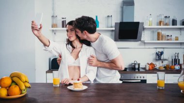 Erkek, düğmesiz gömlekli seksi kadına sarılıyor kahvaltıya yakın akıllı telefondan selfie çekiyor.
