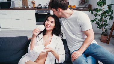 Düğmeleri iliklenmemiş seksi kadın, genç erkek arkadaşının yanındaki kanepede kahve fincanı tutuyor.