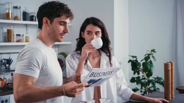 brunette woman in unbuttoned shirt drinking coffee near boyfriend reading newspaper in kitchen clipart