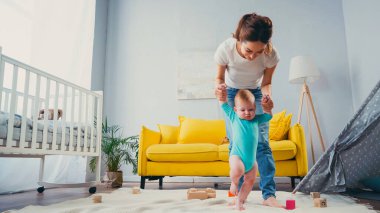 Mutlu anne el ele tutuşuyor ve bebeğe destek oluyor oyuncakların yanında battaniyenin üzerinde yürüyor.