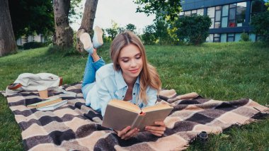 Genç, gülümseyen bir kadın parkta kareli battaniyenin üzerinde uzanırken kitap okuyor. 