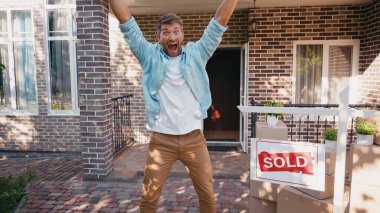 excited man with raised hands and open mouth near board with sold lettering and new house  clipart