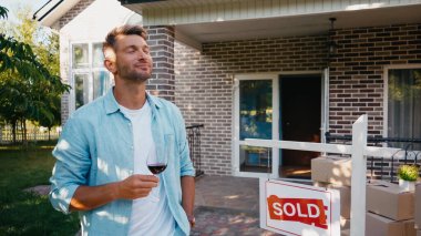 pleased man holding glass of red wine near new house clipart