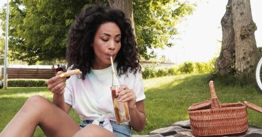 Afro-Amerikan kadın samandan limonata içiyor ve piknik sırasında elinde bir dilim pizza tutuyor. 