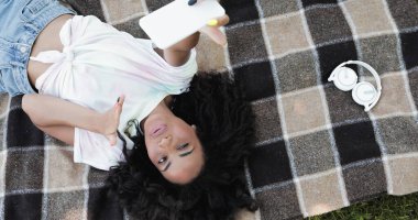 Afro-Amerikalı bir kadının hava öpücüğü gönderdiğini ve piknik battaniyesinde uzanırken akıllı telefondan selfie çektiğini. 
