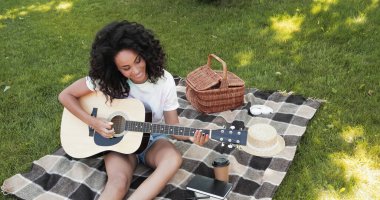 overhead view of cheerful african american woman playing acoustic guitar during picnic in park  clipart