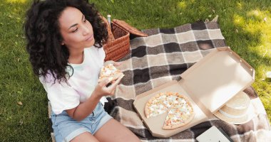 Park 'ta piknik yaparken pizza yiyen kıvırcık saçlı Afro-Amerikan kadının yüksek açılı görüntüsü 
