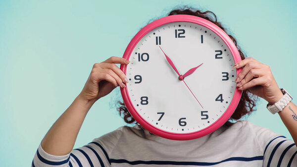 молодая женщина покрывает лицо, держа часы изолированы на синий