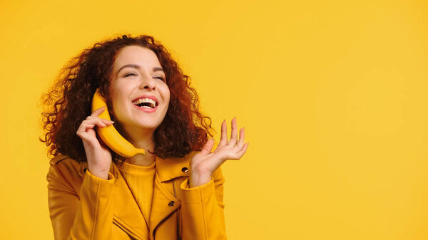 curly woman imitating phone conversation with banana and smiling isolated on yellow