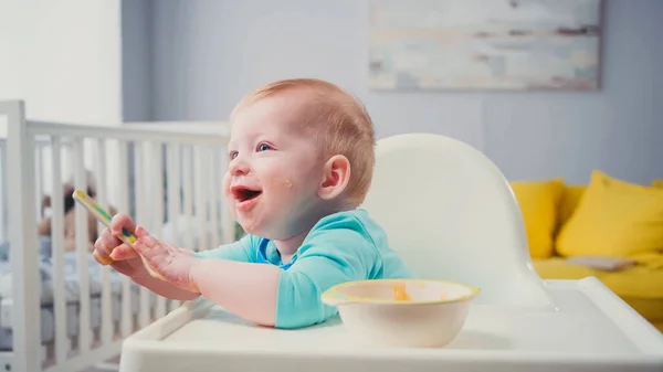 幸せな赤ちゃん青い目をした少年が餌やりの椅子に座ってスプーンを持って — ストック写真