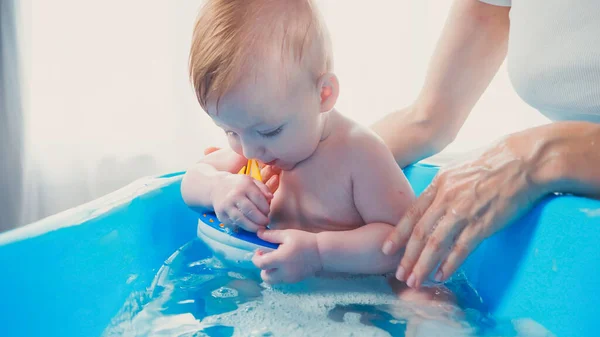 在婴儿澡盆里洗澡的时候 母亲走近婴儿的儿子玩玩具 — 图库照片