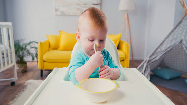 infant boy sitting in feeding chair and holding spoon near bowl 