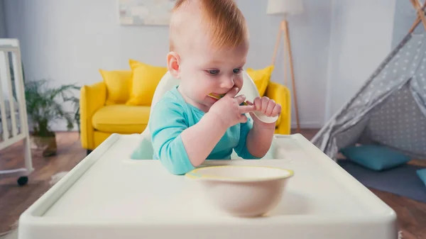 小男孩坐在喂食椅上 在碗边吸勺子 — 图库照片