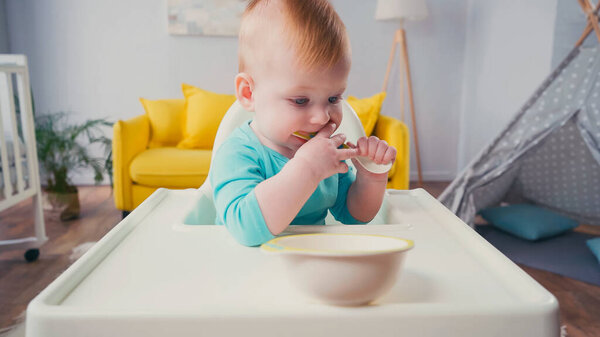 infant boy sitting in feeding chair and sucking spoon near bowl 