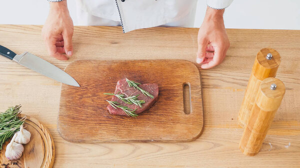 Cropped view of chef standing near beef steak with rosemary branches on chopping board