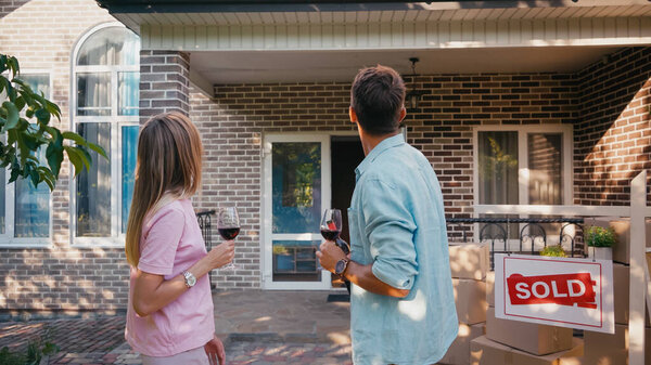 back view of man and woman holding glasses of red wine near board with sold lettering and new house