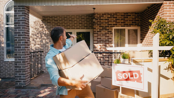 excited man holding boxes and pointing with hand at new house