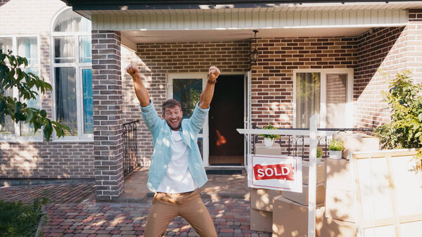 joyful man with raised hands and open mouth near board with sold lettering and new house 