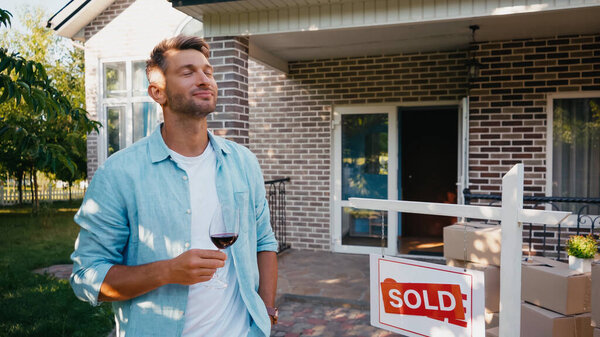 довольный мужчина держит бокал красного вина возле нового дома