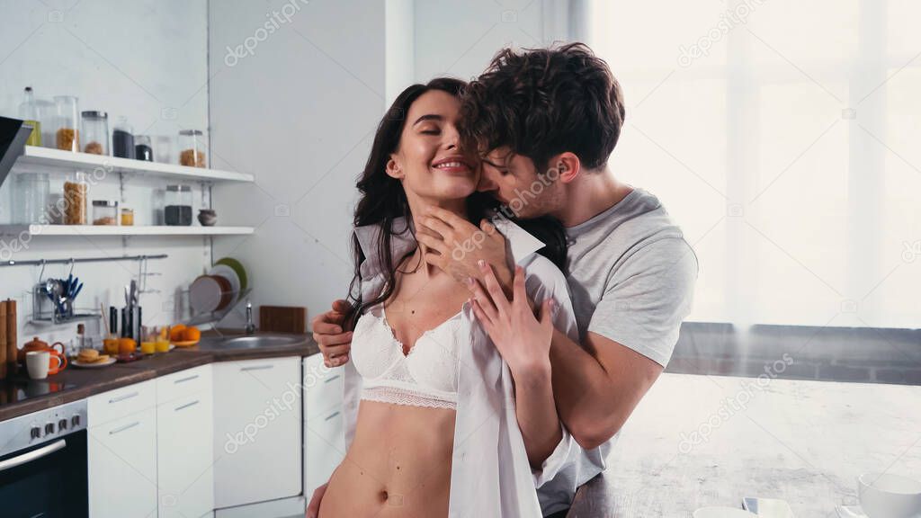 young man embracing sexy, smiling woman in shirt and bra in kitchen