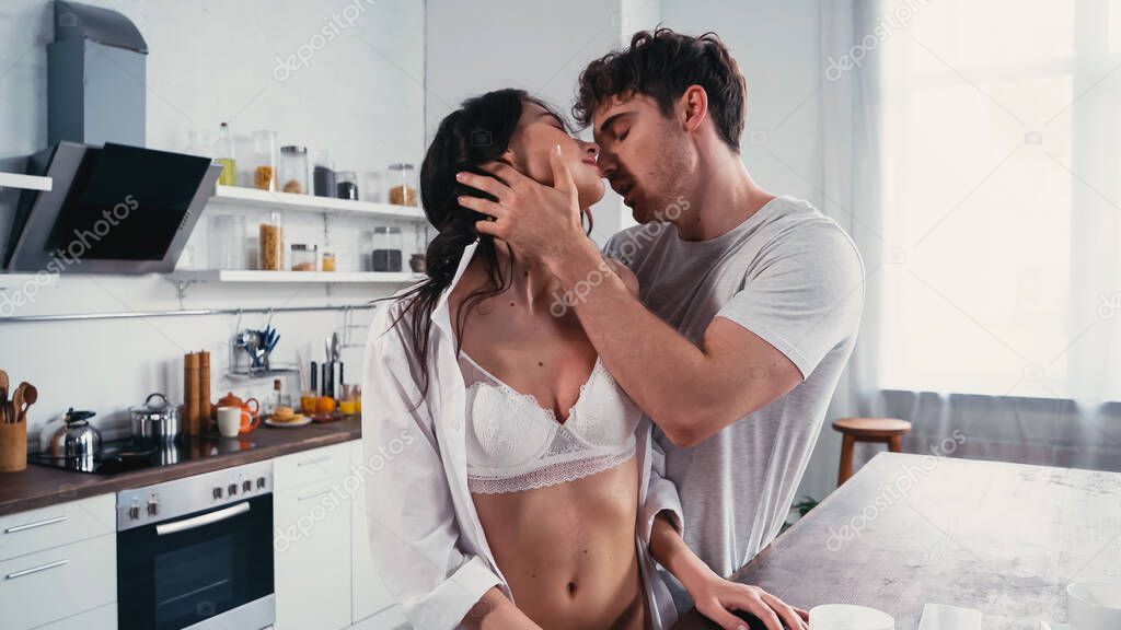 passionate woman in unbuttoned shirt and bra near man kissing her in kitchen