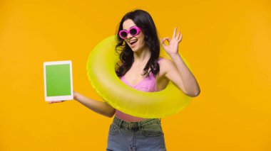 Woman in sunglasses and swim ring showing ok while holding digital tablet with green screen isolated on yellow  clipart