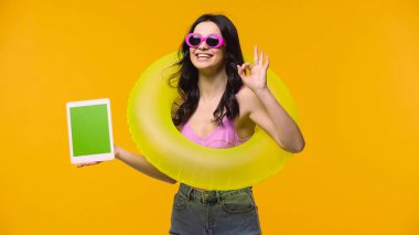 Smiling woman in sunglasses and swimsuit holding digital tablet with green screen and showing okay isolated on yellow  clipart