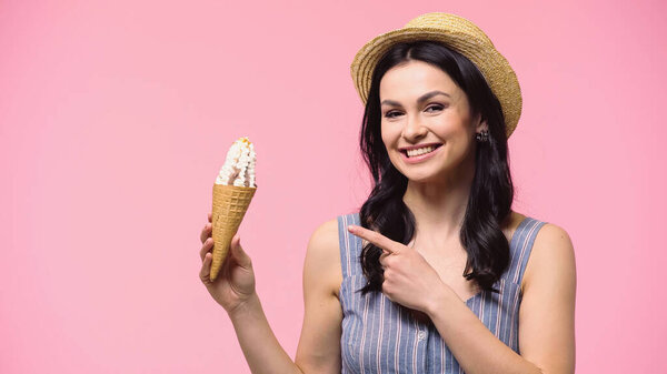 Brunette woman in sun hat pointing at ice cream isolated on pink 