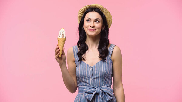 Dreamy woman in straw hat holding ice cream in cone isolated on pink 