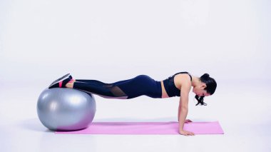Tam boy sporcu kadın, beyaz hasırın üzerinde spor topu ile kalas yapıyor.