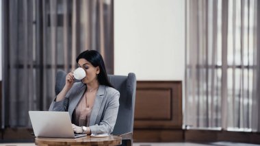 brunette freelancer using laptop and drinking coffee in lobby of restaurant