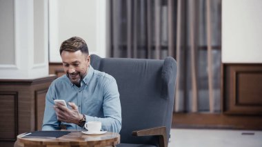 happy businessman using smartphone near cup of coffee and paper folder on table in lobby of restaurant