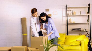 young woman holding book while looking at box and unpacking near boyfriend  clipart