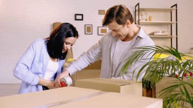 joyful couple using scotch tape while packing box clipart